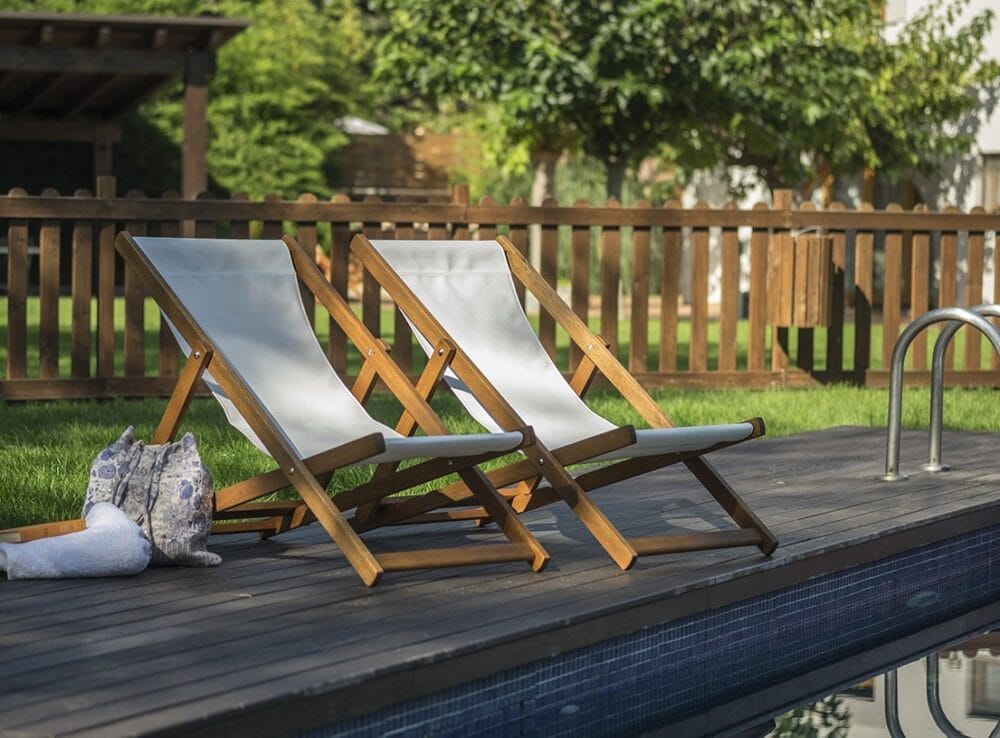 Two mini loungers with a wooden structure and white fabric are seen, the mini loungers are seen in a garden next to a swimming pool