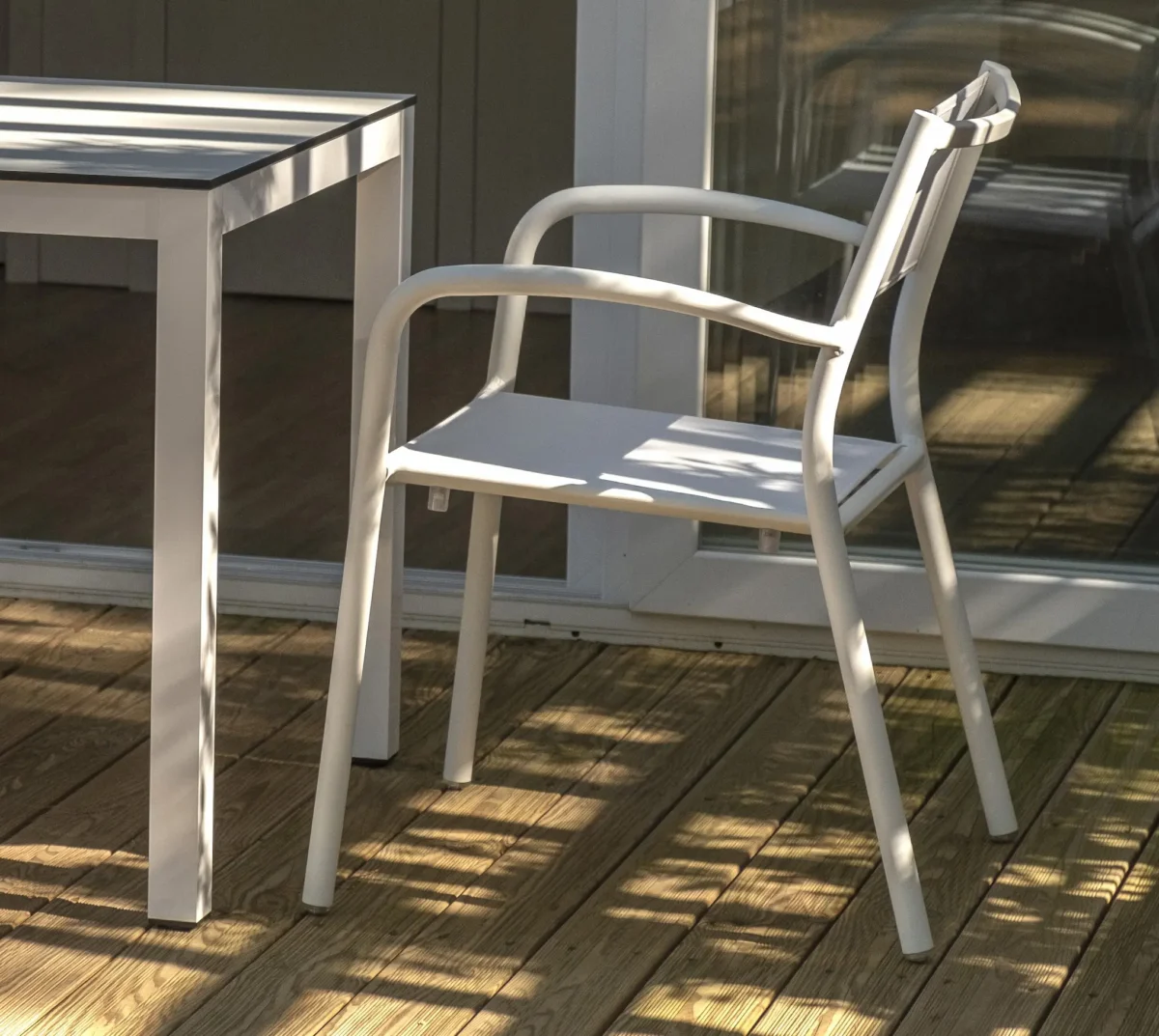 La imagen muestra una silla moderna de color blanco. La silla tiene un diseño minimalista con un respaldo recto y delgado. Sale junto a una mesa en lo que parece ser una terraza