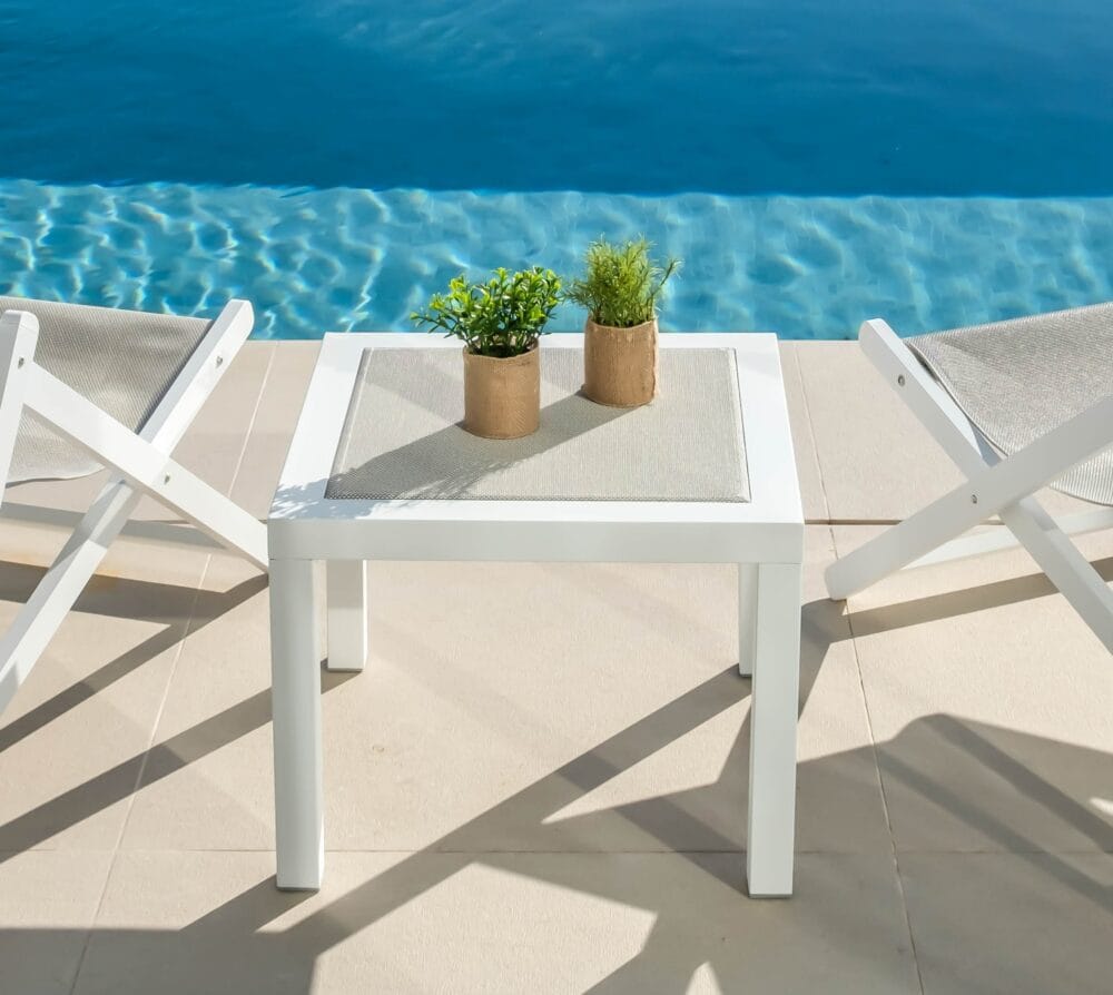 Taula blanca amb el centre del tauler fet de teixit color natural, la taula està al costat de dues minigandules en una piscina