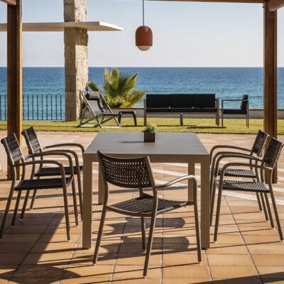 La imagen muestra un conjunto de muebles de terraza que incluye una mesa rectangular con superficie gris y cuatro sillas de diseño moderno con asientos y respaldos trenzados en color negro. El conjunto se encuentra en una terraza pavimentada, bajo una estructura parcialmente cubierta que ofrece vistas al mar. En el fondo, hay un cielo despejado y la silueta de una palmera, lo que añade un ambiente relajante y vacacional al escenario.
