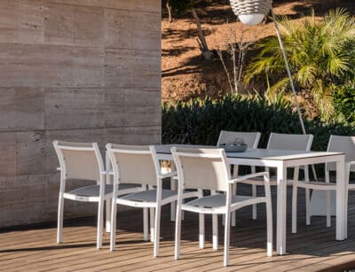 The image shows a set of outdoor furniture consisting of white chairs and a white table. They are placed on a terrace with a wooden floor. The background reveals a natural environment with plants and trees, as well as a concrete wall on the left. The lighting suggests that it is outdoors during the day.