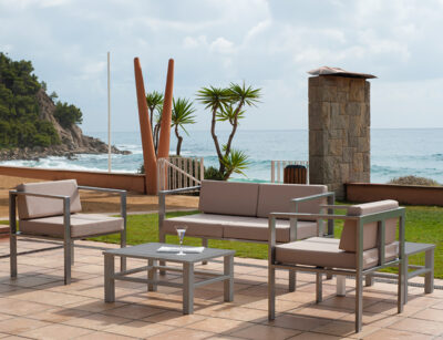 The image shows an outdoor patio setting overlooking a beach. There is a set of modern patio furniture, including a sofa, two armchairs, and a coffee table with a glass on top. In the background, the ocean can be seen.