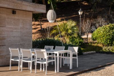 La imagen muestra un conjunto de muebles de exterior compuesto por sillas blancas y una mesa blanca. Están ubicados en una terraza con piso de madera. El fondo revela un entorno natural con plantas y árboles, así como una pared de concreto a la izquierda. La iluminación sugiere que es al aire libre durante el día