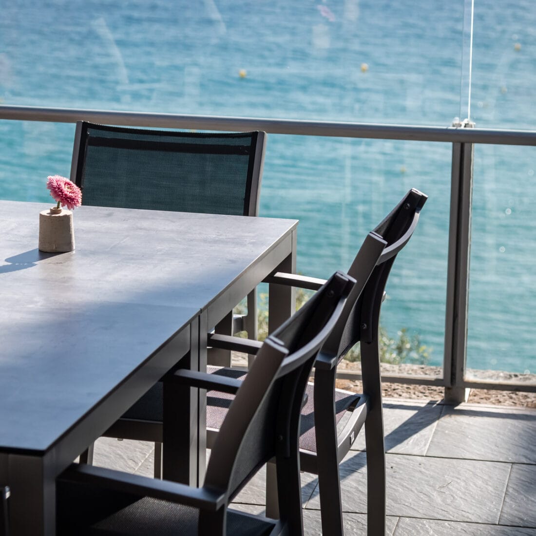La imagen muestra una mesa cuadrada de diseño simple y moderno, con una superficie blanca con tablero HPL y cuatro patas hechas de aluminio . El estilo es minimalista y elegante. El color es gris, con el mar de fondo