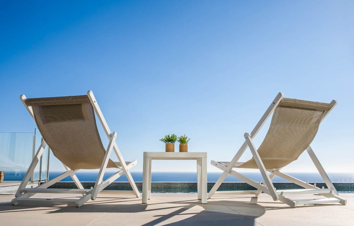 Se muestran dos mini tumbonas plegables de diseño minimalista en color blanco y tela beige, con estructura de madera y asiento de tela resistente, se ve de fondo el cielo azul y una mesita entre las dos mini tumbonas