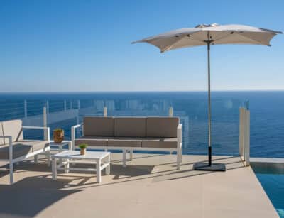 Terrassa moderna amb vista al mar, equipada amb un sofà de 3 places de color beix, un altre sofa però de 2 places, una taula de centre blanca i un para-sol gran obert. La barana de vidre proporciona una vista clar de l'horitzó marí sota un cel clar. Parasol ideal com a para-sol comercial per a restaurant