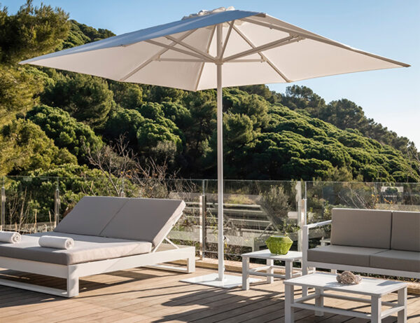 La imatge mostra un ambient de pati exterior amb mobles moderns. Hi ha un gran para-sol blanc que proporciona ombra sobre una gandula reclinable i una zona de seients que inclou un sofà i cadires, tots en tons clars a joc. Ideal per a para-sols comercials per a restaurants.
