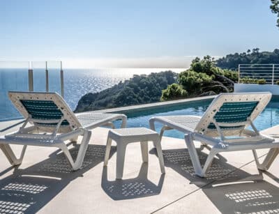 Vista panorámica de dos tumbonas blancas con detalles en azul situadas frente a una piscina infinita que se extiende hacia el mar, rodeada de exuberante vegetación bajo un cielo azul claro