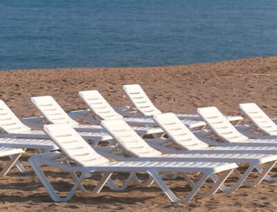 Empty white sun loungers lined up neatly on light sand in front of calm blue waters, evoking calm and rest on a deserted beach.