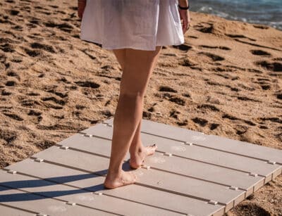 La mitad inferior de una persona de pie en una pasarela de playa en la arena de una playa. Un pie está colocado en la pasarela mientras el otro pie está ligeramente levantado, tocando solo con los dedos de los pies, dando la impresión de movimiento
