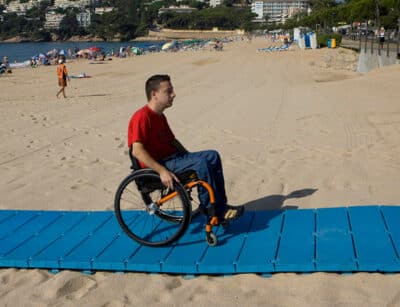 La imagen muestra a una persona utilizando una silla de ruedas sobre una pasarela de playa azul colocada sobre la arena de una playa. La pasarela proporciona un camino estable para la silla de ruedas sobre la arena, facilitando el acceso al área de la playa. En el fondo, hay personas realizando diversas actividades como caminar y tomar el sol.