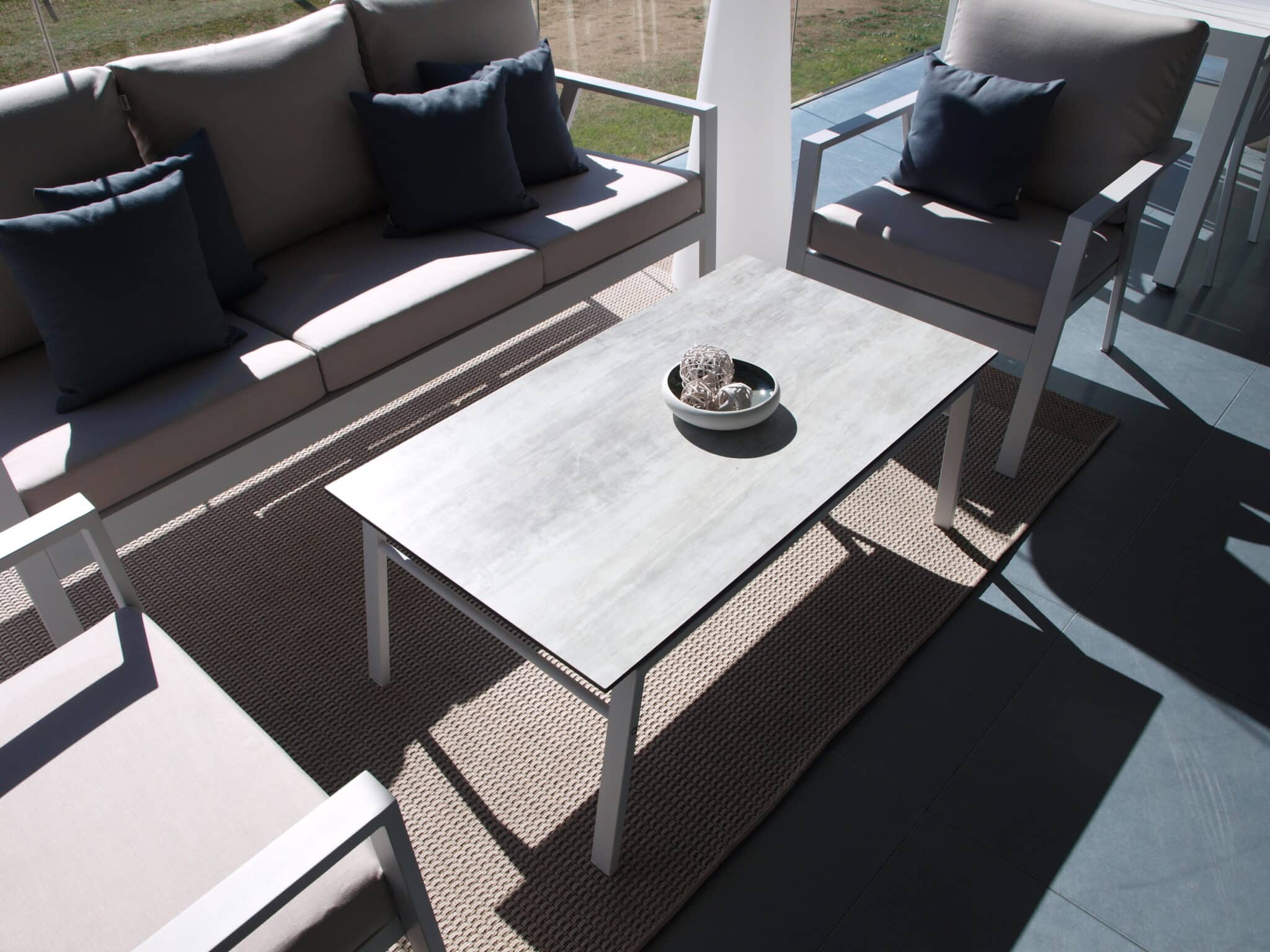 La imagen muestra una mesa de aluminio mate  rectangular de diseño simple y moderno junto a un sofá creando un conjunto, con una superficie gris con tablero HPL y cuatro patas hechas de aluminio . El estilo es minimalista y elegante