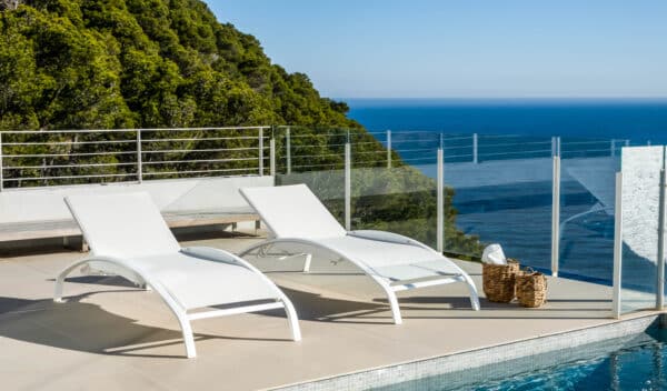 The image shows two modern white sun loungers with adjustable backrests and a sleek, curved design. The sun loungers are placed against a light, simple background, highlighting their clean lines and contemporary style. They are part of the Balliu range of aluminium sun loungers for solariums.