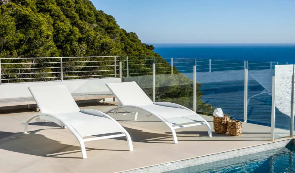 The image shows two modern white sun loungers with adjustable backrests and a sleek, curved design. The sun loungers are placed against a light, simple background, highlighting their clean lines and contemporary style. They are part of the Balliu range of aluminium sun loungers for solariums.