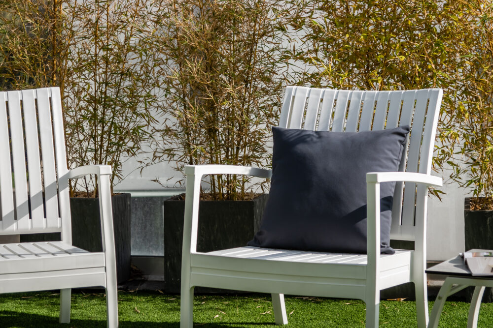 In the image you can see two white armchairs, with a modern and elegant design, with a black cushion on top of the armchair, and in the background you can see vegetation.